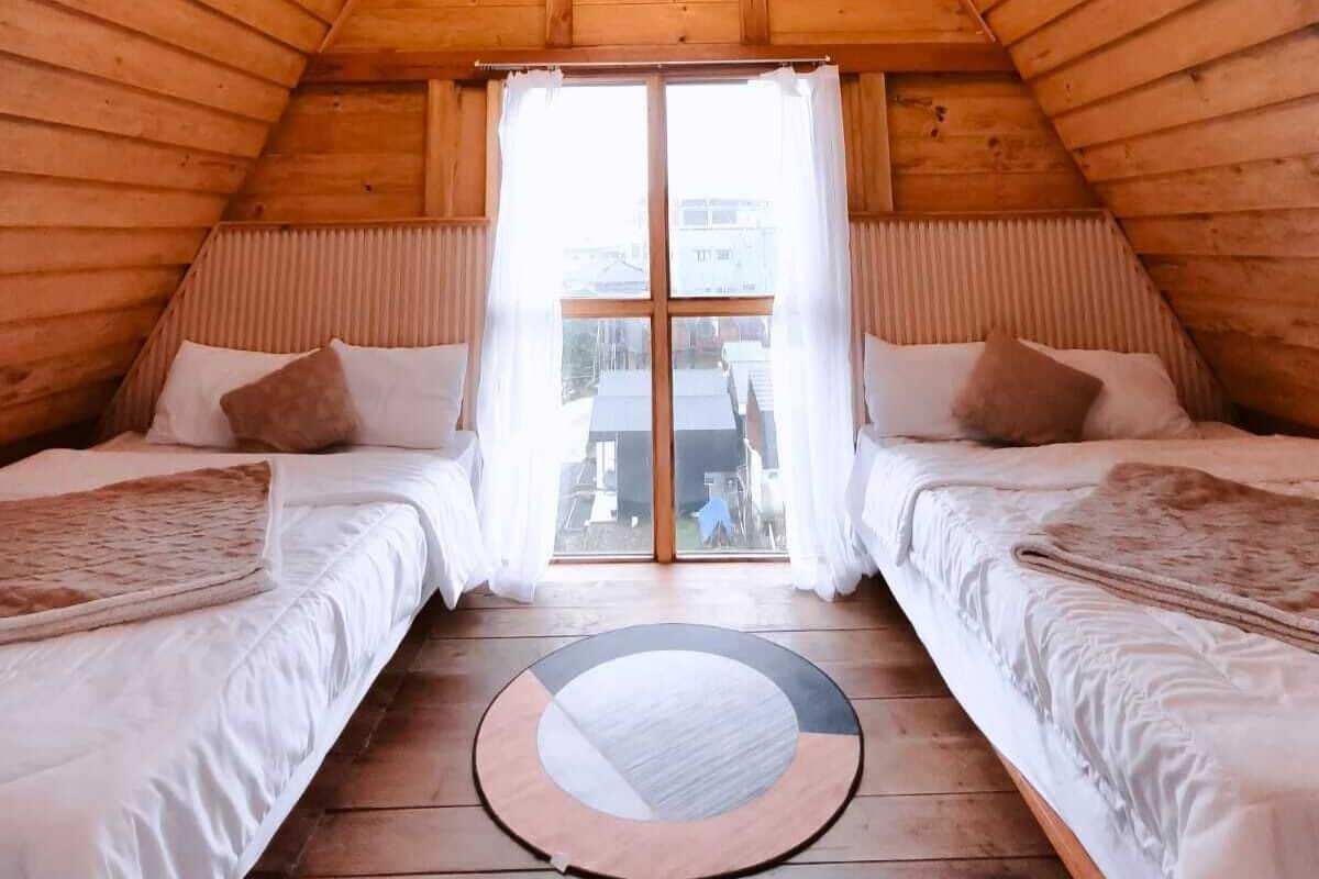 A cozy attic bedroom with wooden walls, a sloped ceiling, two beds, a circular rug, and a window with curtains.