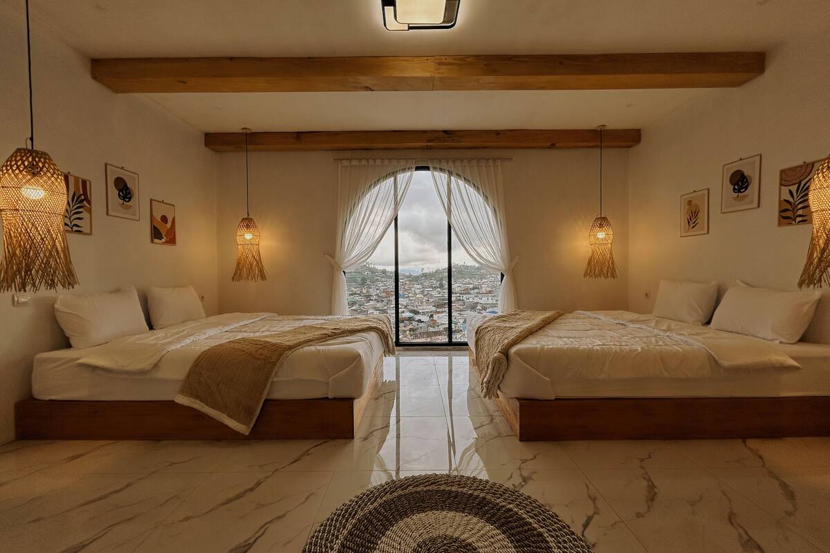 A room with two beds, a large window with a view, and hanging lamps.