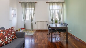 Um quarto com um sofá cinzento, uma mesa de jantar com cadeiras e duas janelas com cortinas.