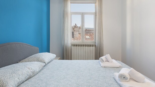 Quarto com cama, janela com cortinas e vista para edifícios.