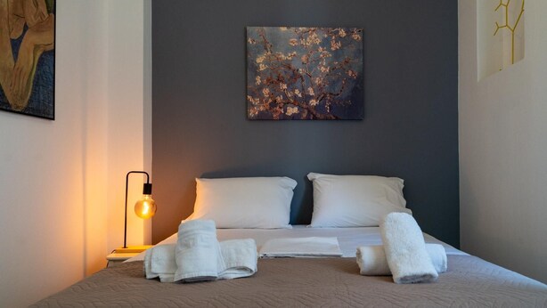 A neatly made bed with white linens and two pillows, a wall-mounted painting of a tree branch, and a bedside lamp with a yellow light.