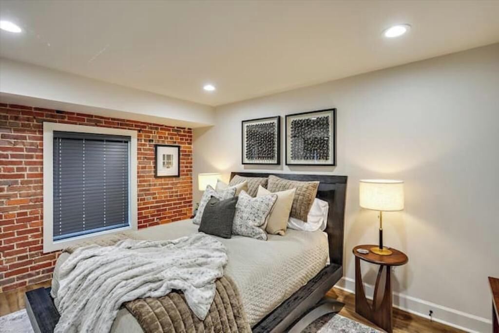 Un dormitorio con una pared de ladrillo decorativa, una cama con muchas almohadas, una lámpara de noche y dos cuadros enmarcados en la pared.