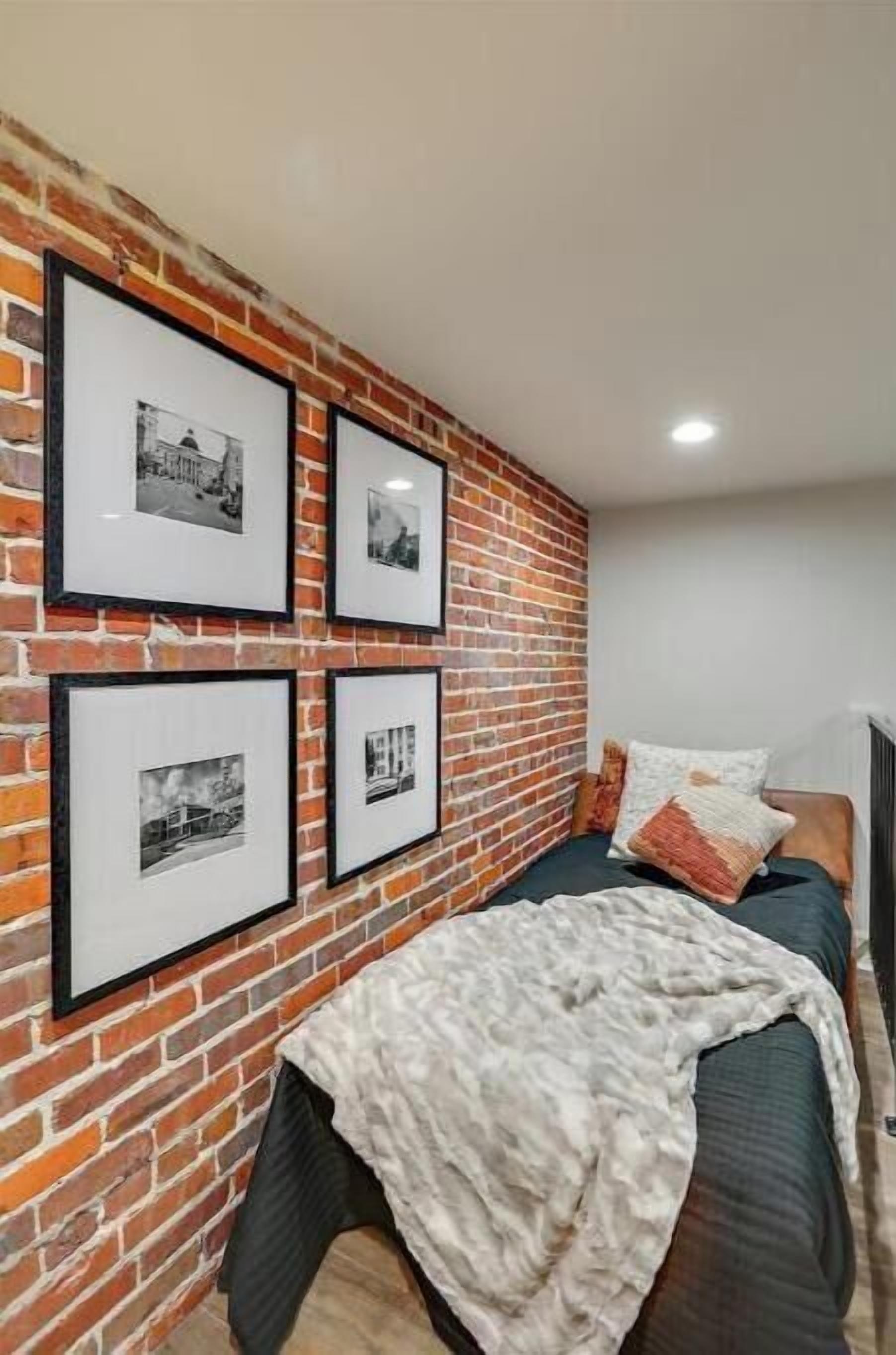 A room with a brick wall, a bed with a white blanket, and four framed pictures.
