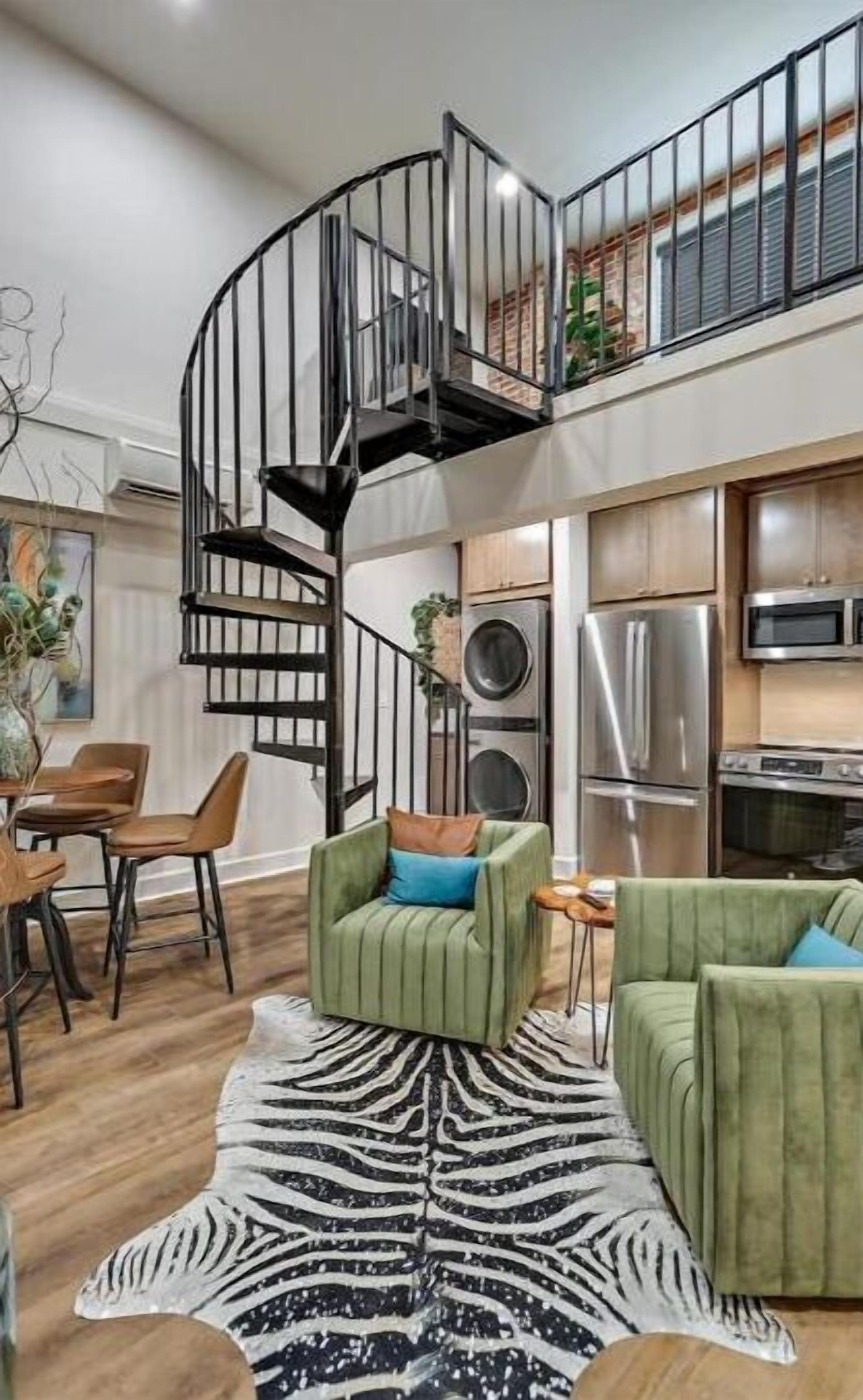 A modern living room with a spiral staircase, green armchairs, a zebra-patterned rug, and a kitchen area with stainless steel appliances.