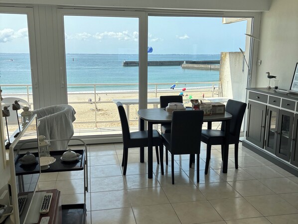 A room with a large window overlooking a beach, a dining table with chairs, a kitchen area with cabinets, and a small shelf with decorative items.