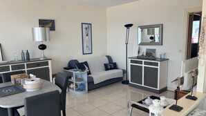 A modern living room with a sofa, a dining table, and a kitchen area with a fridge and cabinets.
