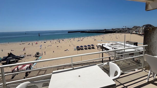 A balcony with a view of a beach, a dining area with tables and chairs, and a clear blue sky.