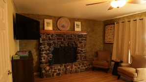 A living room with a stone fireplace, a flat-screen TV mounted above it, a wooden floor, and two leather armchairs.
