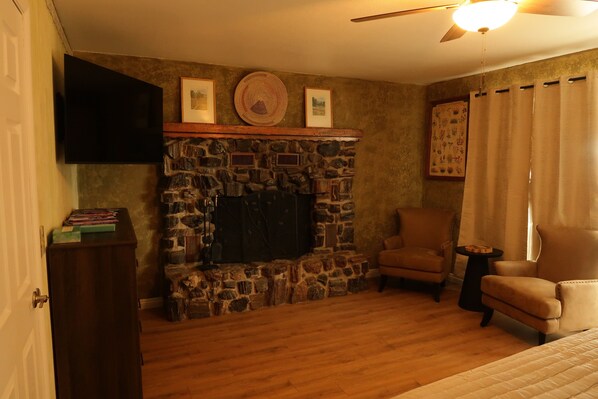 A living room with a stone fireplace, a flat-screen TV mounted above it, a wooden floor, and two leather armchairs.