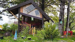 A rustic wooden cabin with a large wheelhouse, surrounded by greenery and rocks.