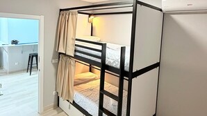 Uma cama beliche com uma escrivaninha e cadeira num quarto com uma cozinha compacta ao fundo.