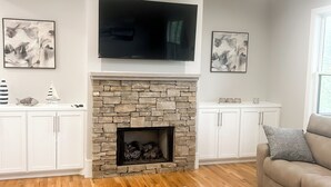 A living room with a stone fireplace, a flat-screen TV mounted above it, and a ceiling fan.