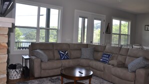 A living room with a beige sectional sofa, two windows with a view of greenery, a fireplace, and a ceiling fan.