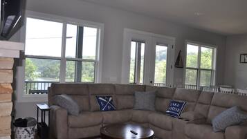 Un salon avec un canapé d’angle beige, deux fenêtres donnant sur un paysage verdoyant, une cheminée et un ventilateur de plafond.