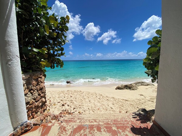 Vista de una playa con agua turquesa cristalina, una orilla arenosa y un muro de piedra.