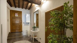 A hallway with a wooden door, a white wall-mounted mirror, a small white table with a potted plant, and a large potted plant to the right.