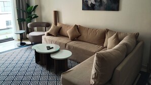 Un salon avec un canapé beige, deux tables d’appoint, un grand tableau représentant des fleurs, une plante en pot et une vue sur l’extérieur par une fenêtre ornée de rideaux.