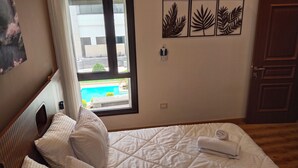 Une chambre d’hôtel avec un lit, une fenêtre donnant sur une piscine, une porte et des œuvres d’art mural.