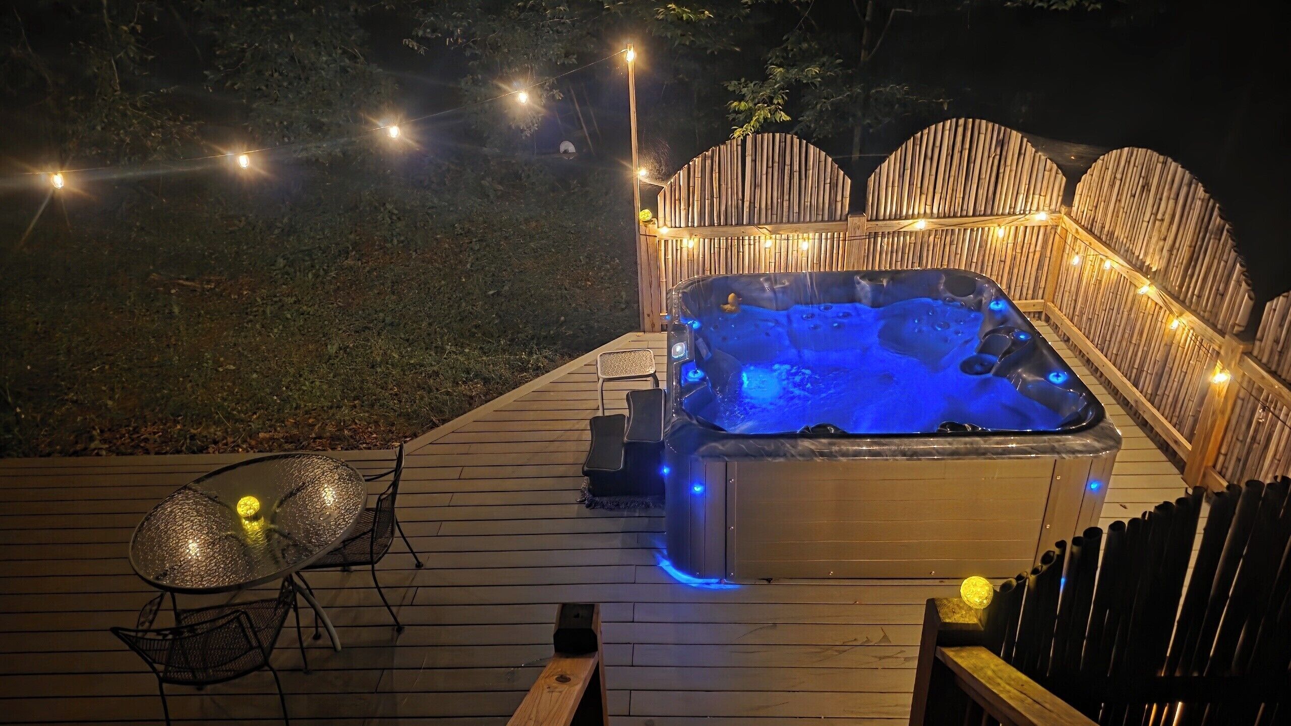 Un bain à remous sur une terrasse en bois, agrémentée de guirlandes lumineuses, avec une table en verre et des chaises.