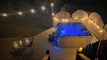 Un bain à remous sur une terrasse en bois, agrémentée de guirlandes lumineuses, avec une table en verre et des chaises.
