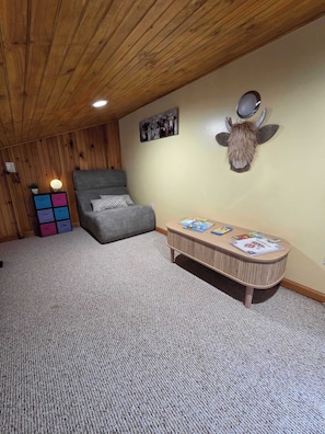 A room with a wooden ceiling, a gray sofa, a small table with magazines, a wall-mounted animal head decoration, and a colorful storage unit.