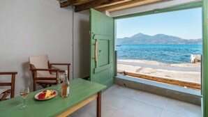 Een kamer met een groene tafel, twee stoelen, een fles wijn en een schaal fruit. Een groot raam biedt uitzicht op zee en bergen.