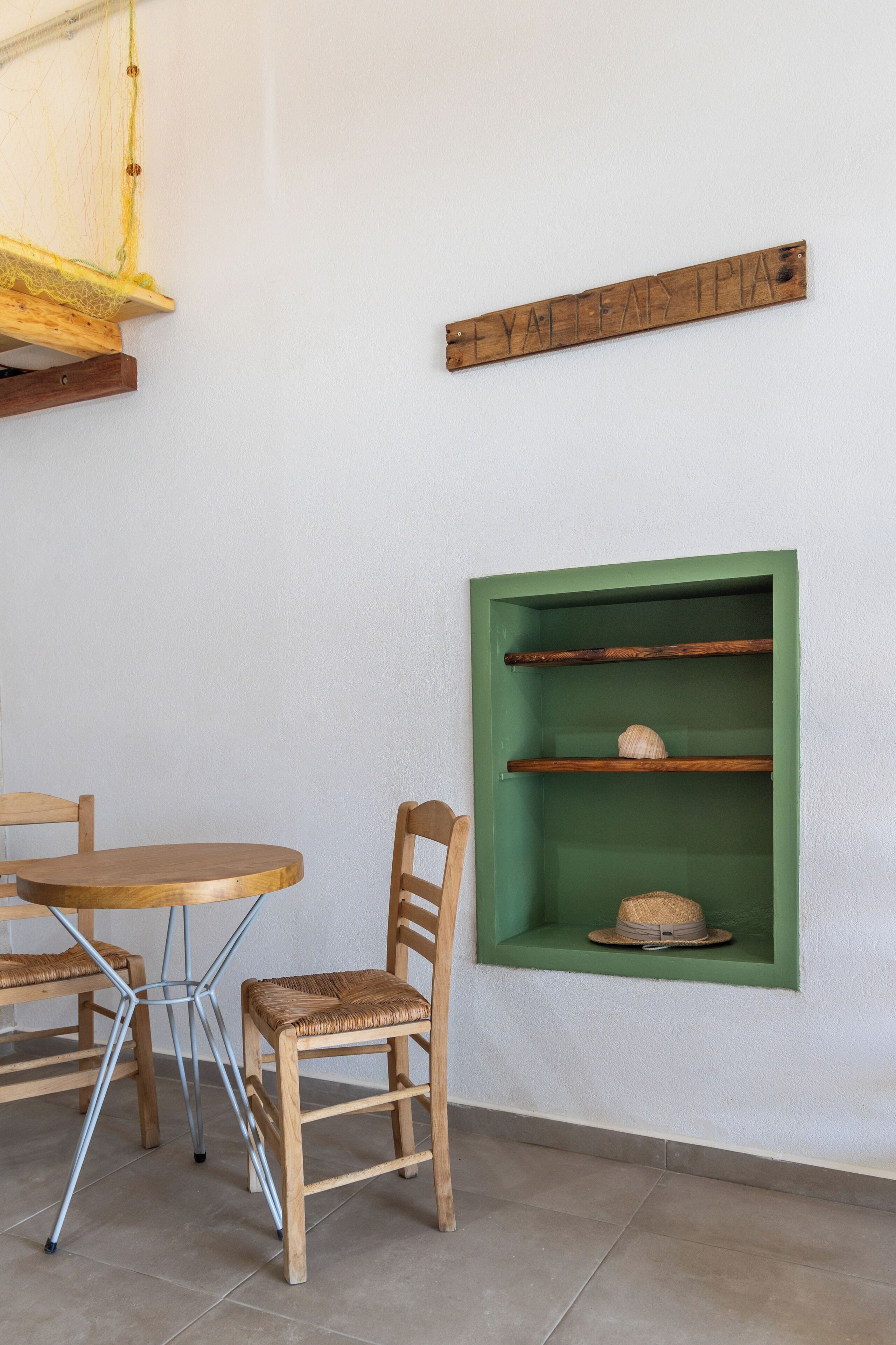 Una habitación con una mesa redonda de madera y dos sillas de mimbre, un estante verde con un sombrero, y un letrero de madera en la pared.