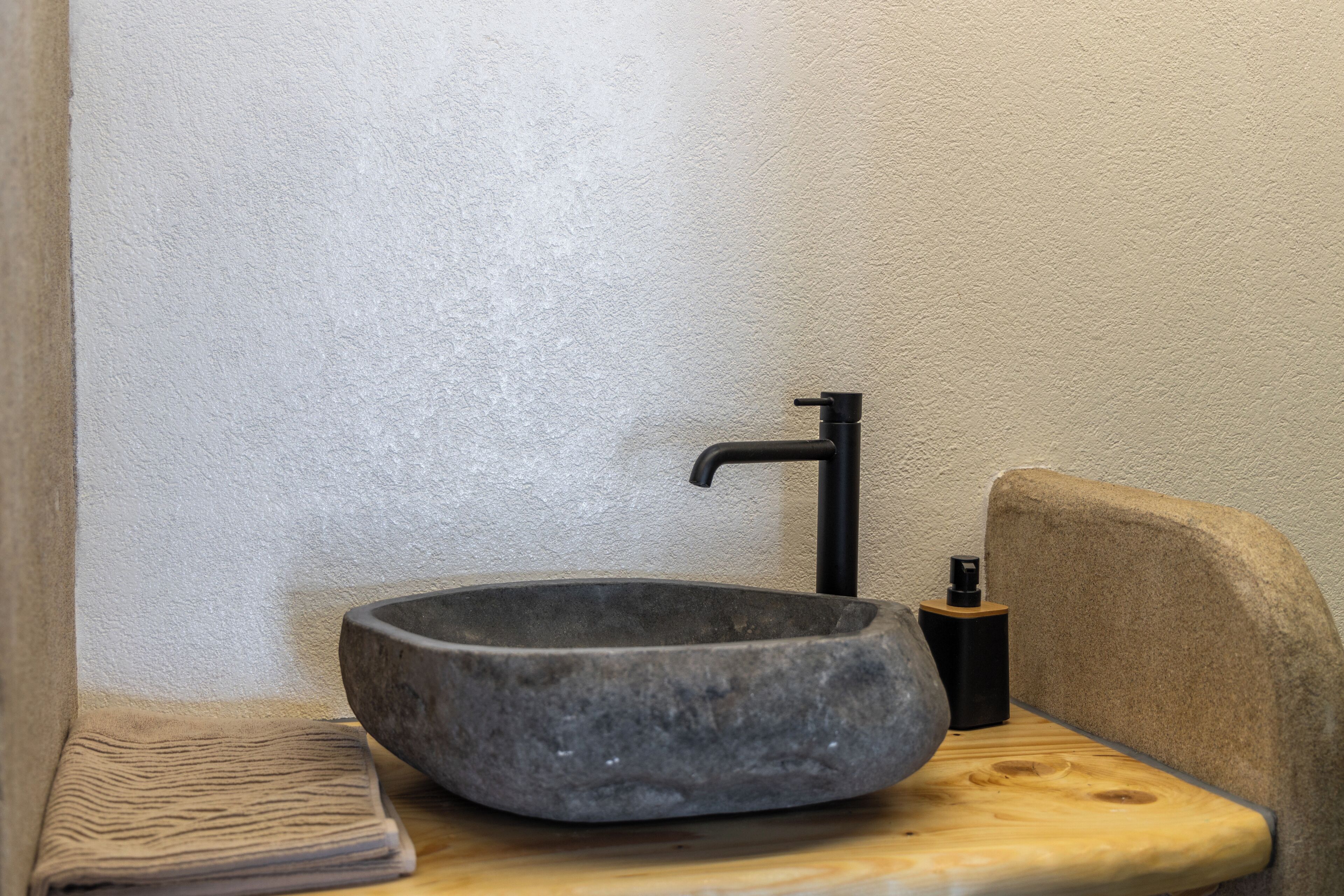 Un évier en pierre avec un robinet noir, placé dans un coin d’une pièce dont le mur est texturé et où se trouve un banc en bois.
