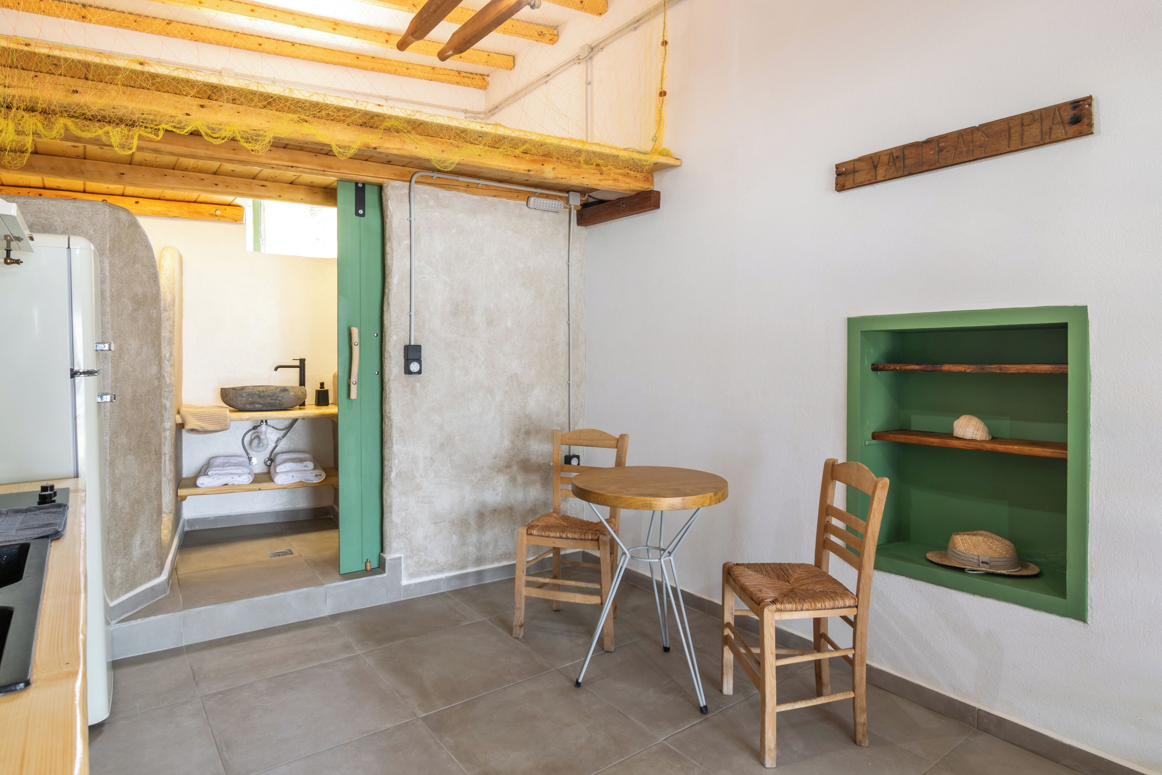 Una stanza con soffitto in legno, uno scaffale verde con cappelli, un tavolo e sedie in legno, e una porta che conduce a un'altra stanza.