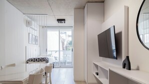 A modern, minimalist room with a white dining table, chairs, a flat-screen TV mounted on a white wall, and a large window offering a view of the outdoors. - BT Homes - Palermo, Oro  (Buenos Aires)
