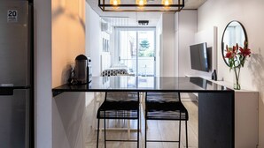 A modern kitchen with a stainless steel refrigerator, a glass island with two black bar stools, a coffee machine, and a flat-screen TV mounted on the wall. - BT Homes - Palermo, Oro  (Buenos Aires)
