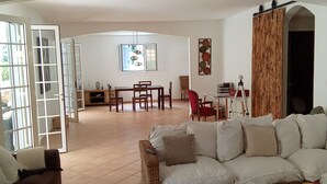 A spacious living area with a sofa, a wooden coffee table, and a dining table with chairs. There is a sliding door leading to an outdoor area.