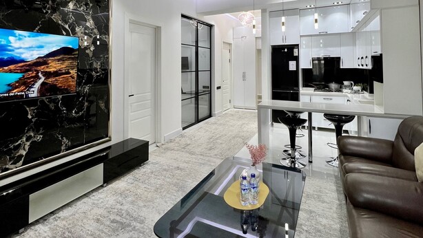 A modern living room with a large flat-screen TV, a glass coffee table, a brown leather sofa, and a kitchen in the background. - Bibigon 31 (Tashkent)