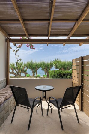 A covered patio with a table and two chairs, overlooking a garden with trees and a view of the sea.