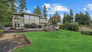 A two-story house with a well-maintained lawn, a wooden deck, and a variety of trees and shrubs in the background.