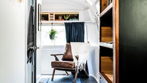 Interior de autocaravana moderno y compacto, con techo de madera, zona de estar con sillón de cuero marrón, una mesita y ventana con vistas.