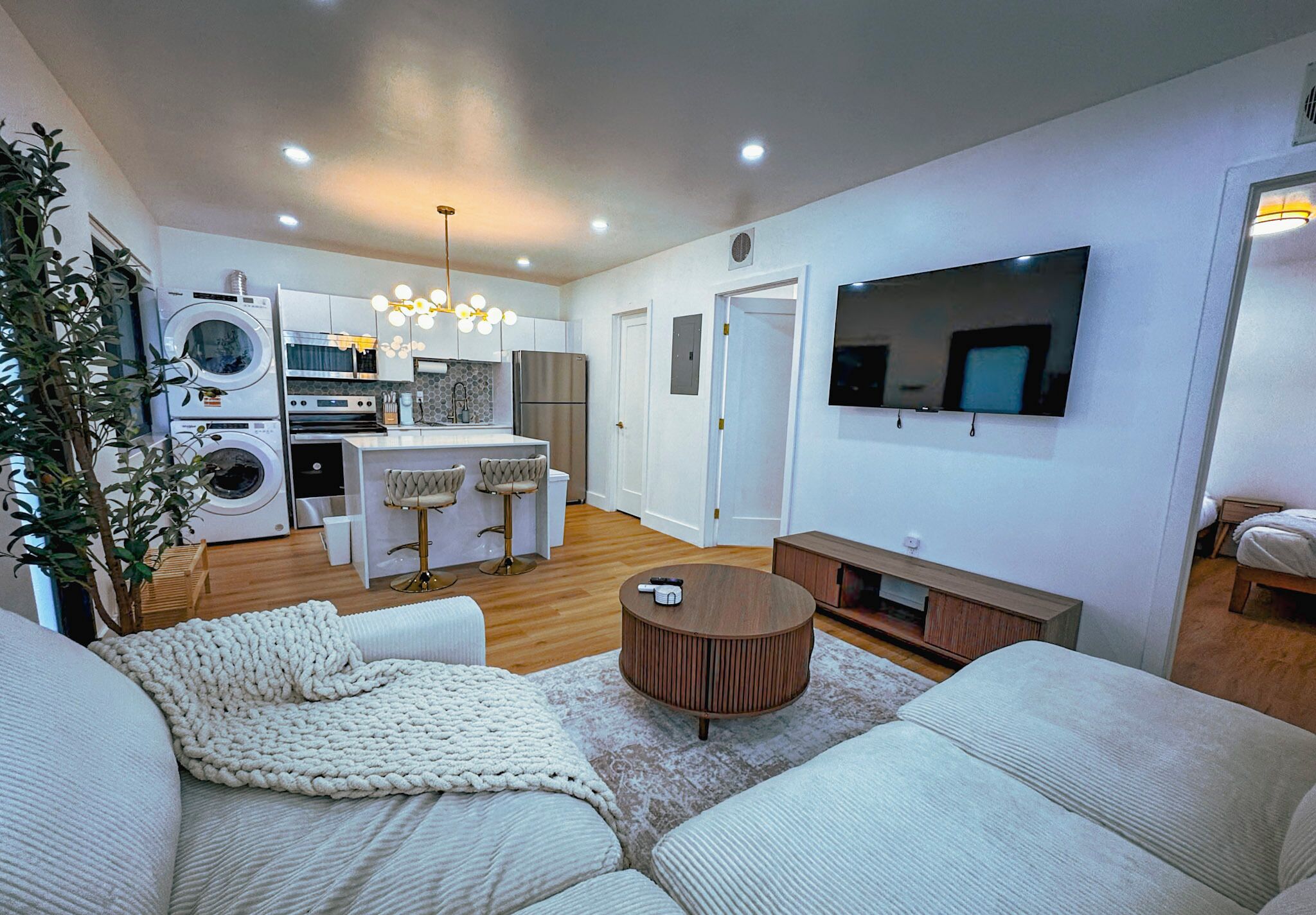 A modern living room with a sofa, a coffee table, and a TV. There is a kitchen area with appliances and a dining table.