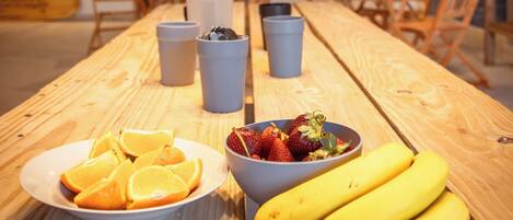 A long wooden table with a plate of sliced oranges, a bowl of strawberries, and three bananas.