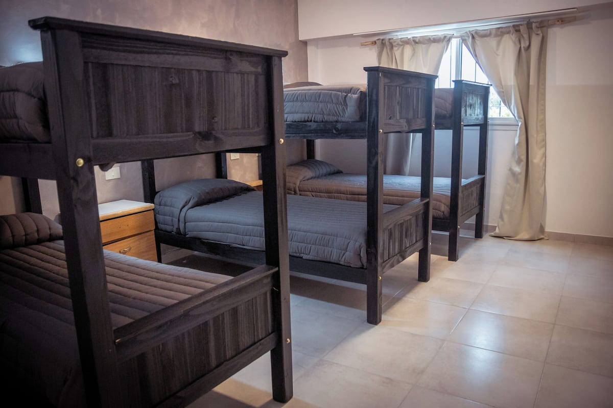 Um dormitório com beliches, uma mesinha de cabeceira de madeira e uma janela com cortinas.
