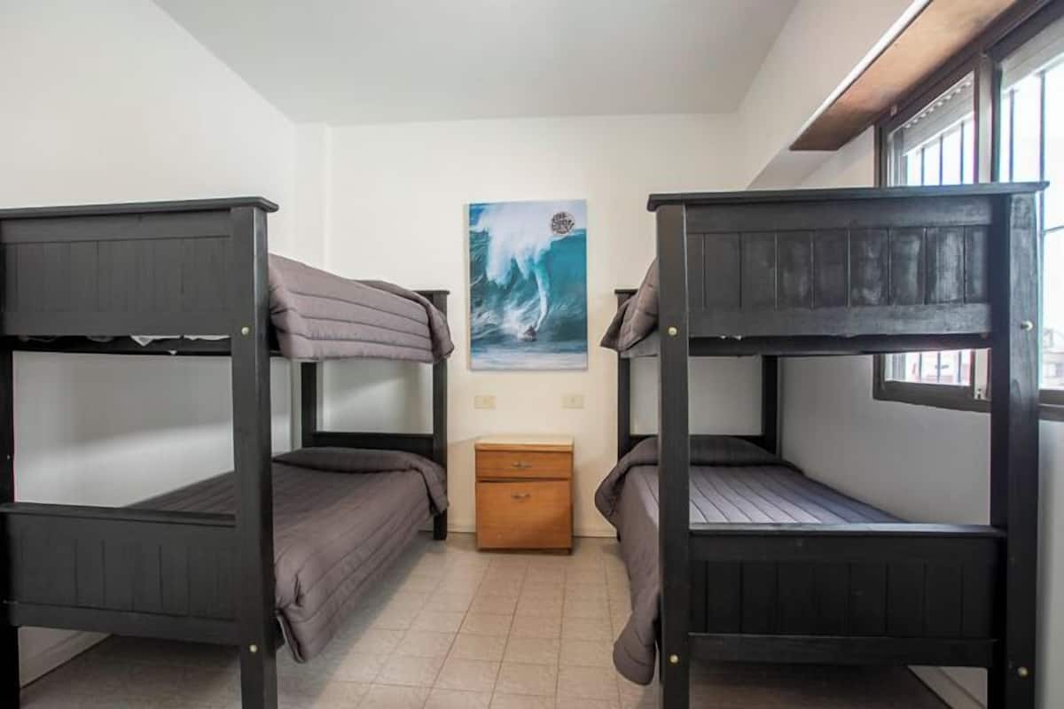 Quarto em dormitório com beliches, uma pequena mesa de cabeceira de madeira e um quadro de uma onda na parede.