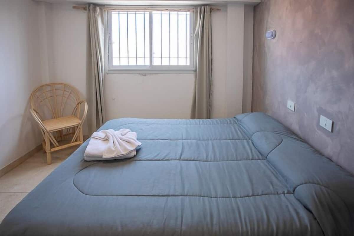 Uma cama de solteiro com colchão cinzento e toalha branca dobrada, uma cadeira de madeira e uma janela com grades verticais.