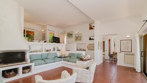 Amplia sala de estar con chimenea, sofá modular y una mesita de centro. Hay estanterías con objetos decorativos y un ventanal que proporciona luz natural.