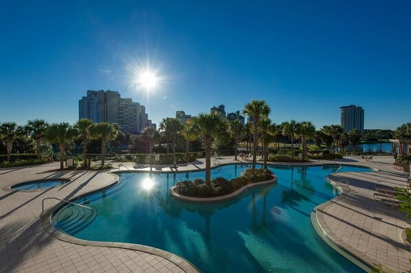 A large outdoor swimming pool with a sunlit sky, surrounded by palm trees and a cityscape in the background.