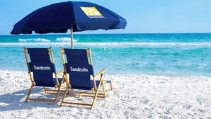 Zwei Strandstühle mit 'Sandestin'-Branding und ein passender Sonnenschirm an einem Sandstrand, im Hintergrund der Ozean.
