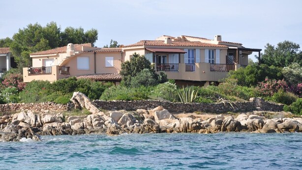 A coastal property with a large house featuring a balcony, surrounded by lush greenery and a rocky shoreline.