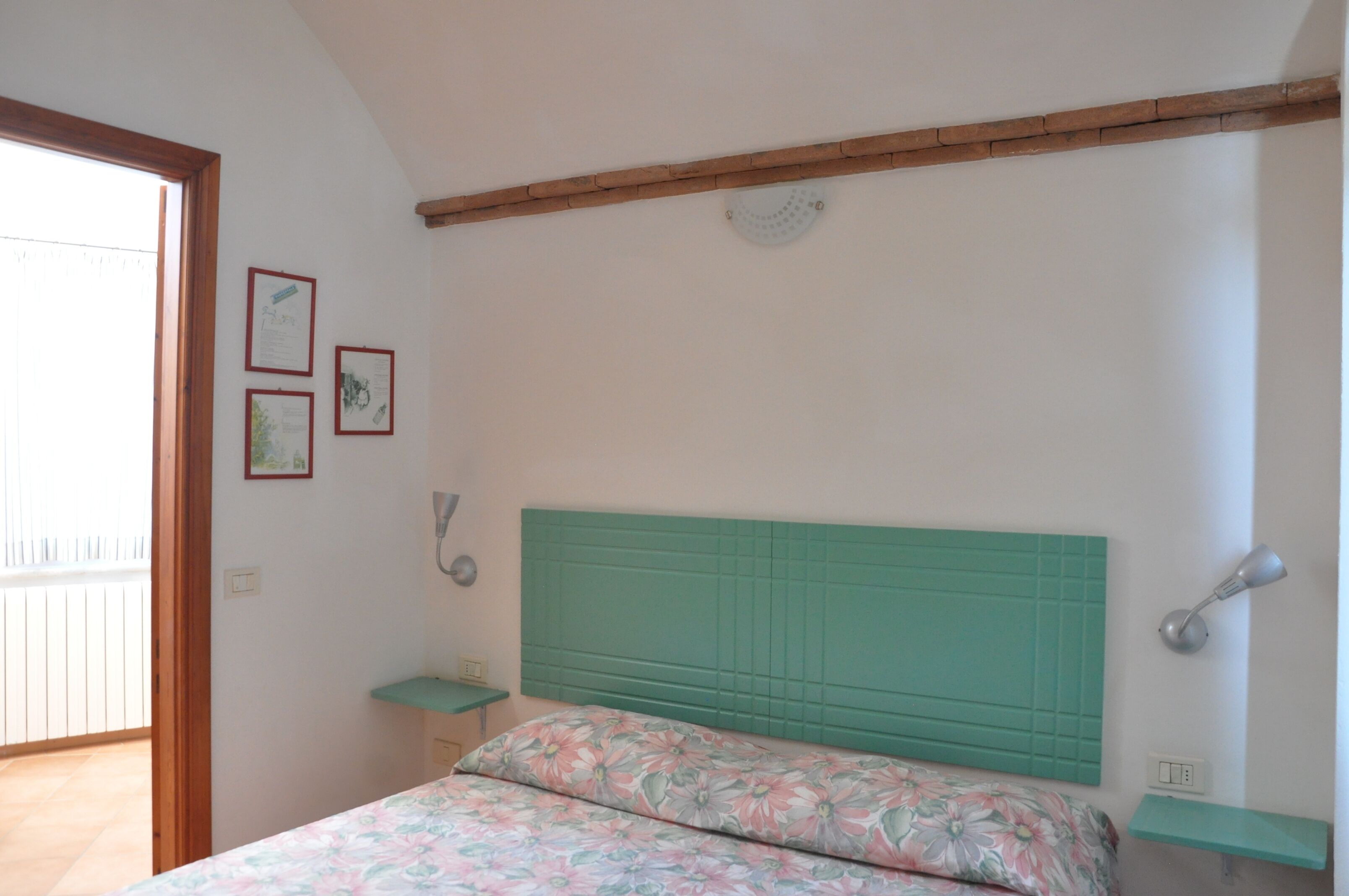 Een slaapkamer met een groen hoofdeinde, een bloemenbedekker, een klein nachtkastje en een wandlamp.