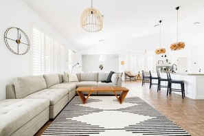 A modern living room with a sectional sofa, a coffee table, and a dining area with chairs and a kitchen in the background. - Village House: Pool, Spa, Golf! (Queen Creek)