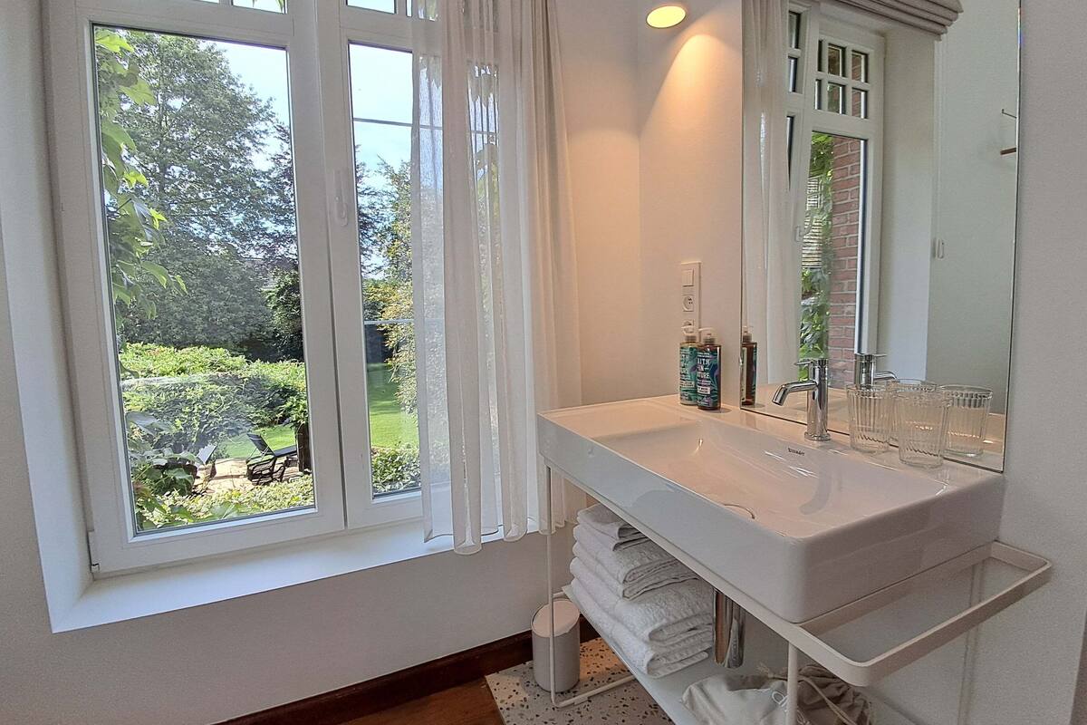 Une salle de bain avec une grande fenêtre, un lavabo blanc, un miroir et un porte-serviettes.
