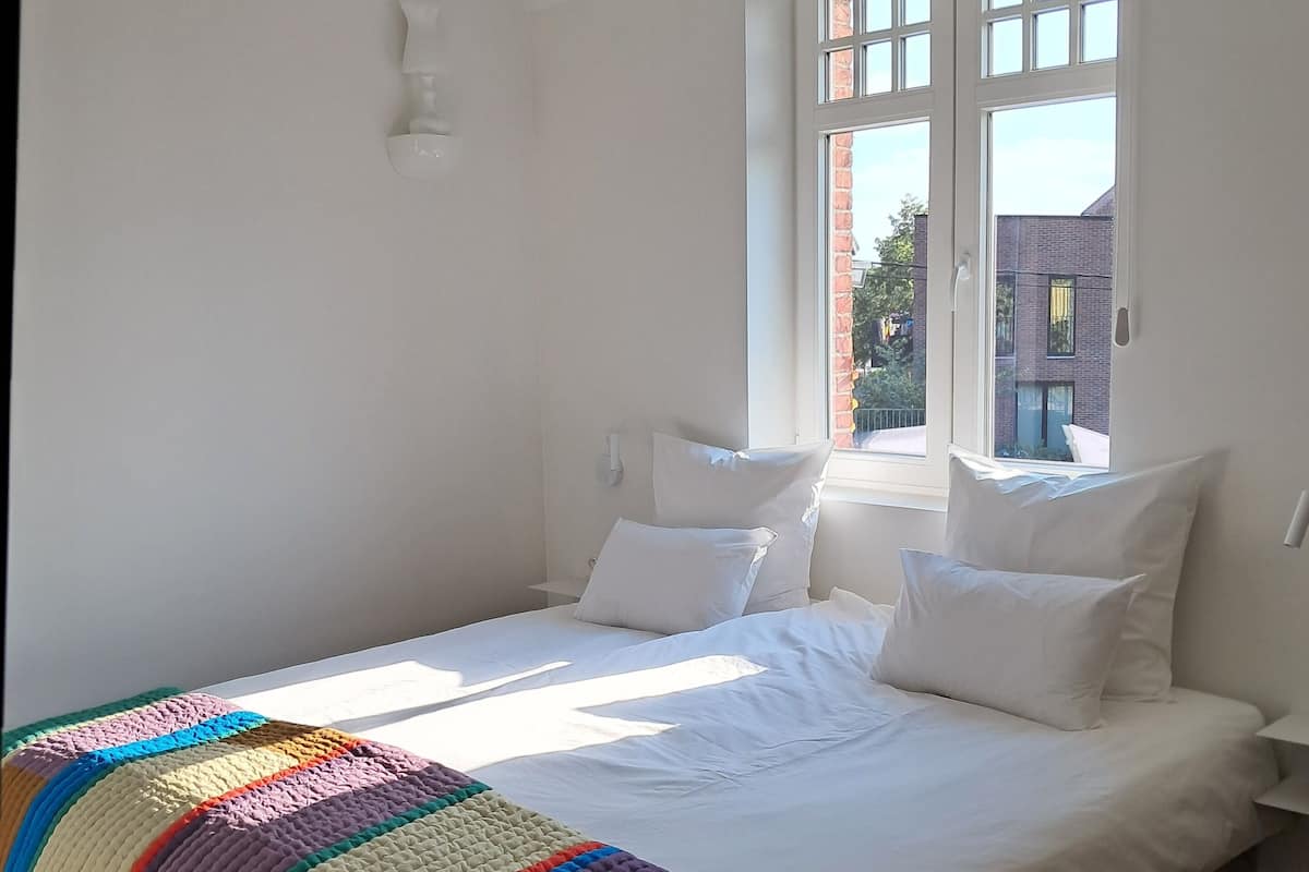 Une chambre avec un grand lit, du linge de lit blanc, une couverture colorée, une fenêtre donnant sur des immeubles et un plancher en bois.