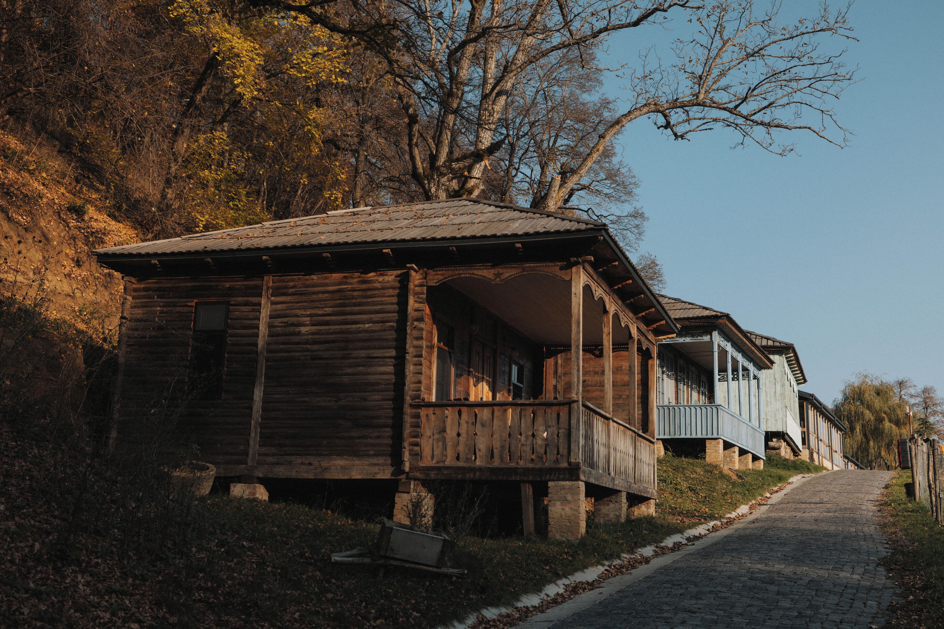 A rustic wooden house with a porch, situated on a cobblestone path, surrounded by trees and a clear sky.
